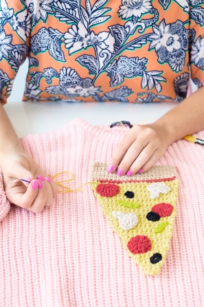 Someone sewing a slice of pizza onto a pink sweater