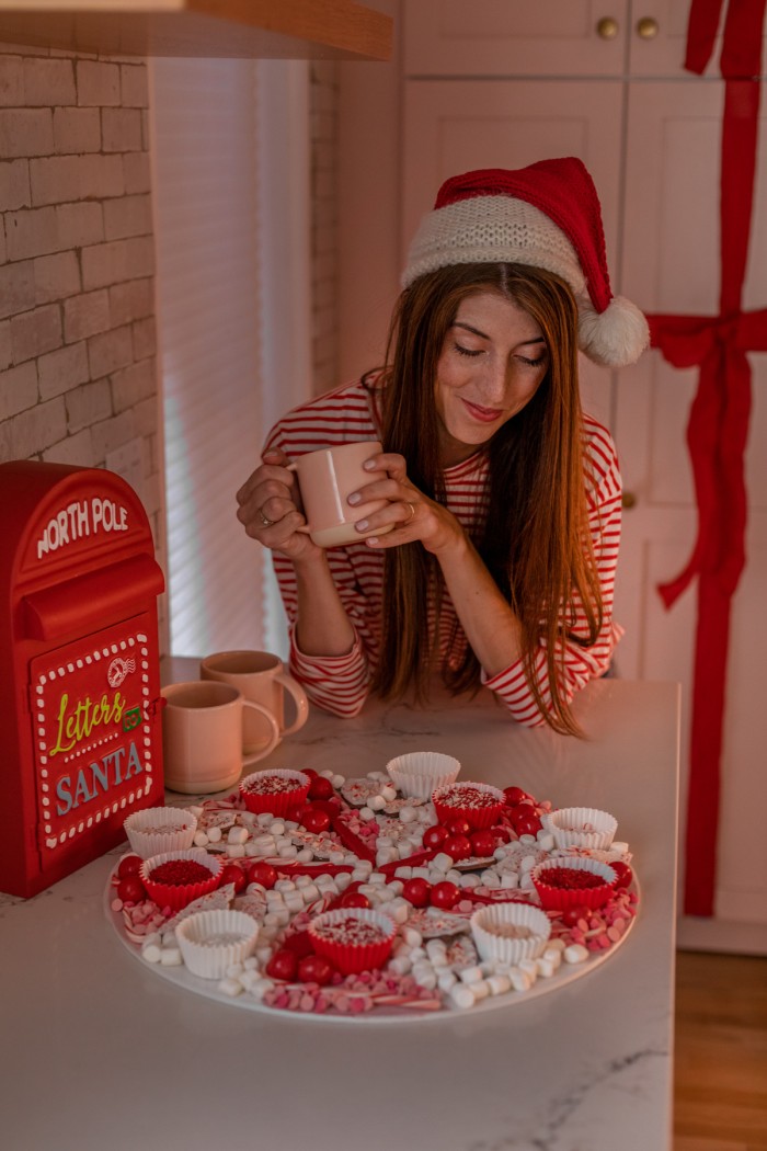 Hot Chocolate Toppings Christmas hot chocolate board with a woman in the background holding a mug and wearing a Santa hat.
