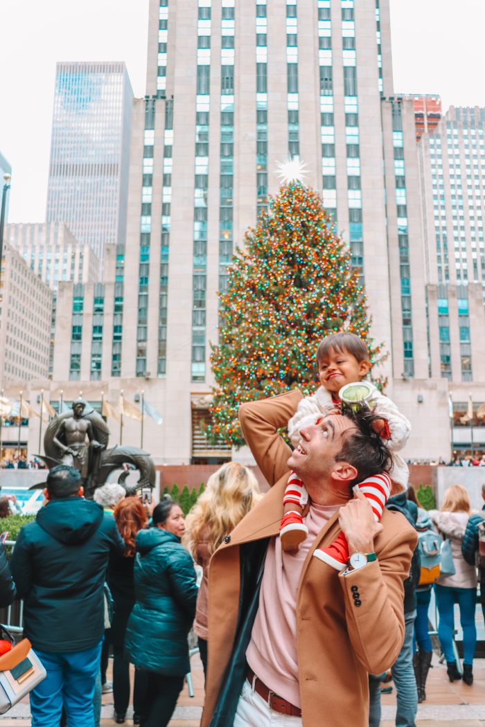 Holiday Bucket List Ideas Man with a child on his shoulders standing in front of the Christmas tree at Rockefeller center.