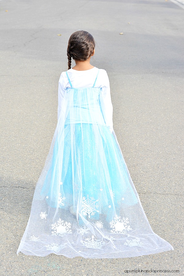 Elsa Christmas Costume Girl standing with her back to the camera, dressed with a blue dress and long white veil with snowflakes on it.