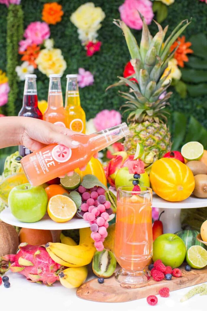 Moana Fruit Juice Hand holding a bottle of soda and pouring it into a glass surrounded by tropical fruits.